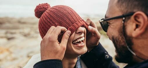 Les visages d'un homme et d'une femme sur une plage sont en avant plan de l'image, Elle rit parce qu'il lui descend son bonnet sur les yeux.