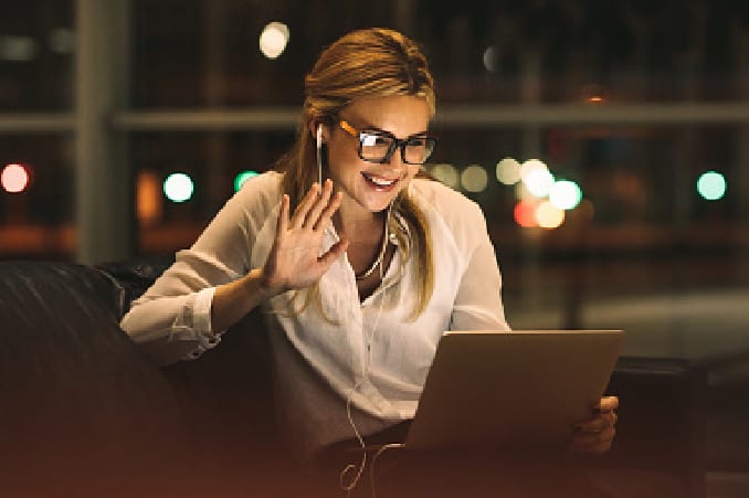 Une femme dans son canapé regarde son ordinateur et fait coucou de la main à son écran en souriant